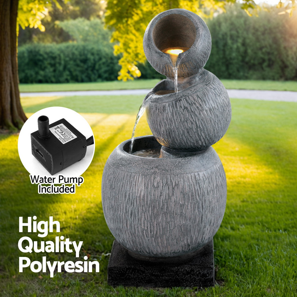 A three-tier bowls cascading water fountain feature with LED lights, made of polyresin, in a grey colour with black base, insert image of the water pump included, all in a garden setting with trees in the background.