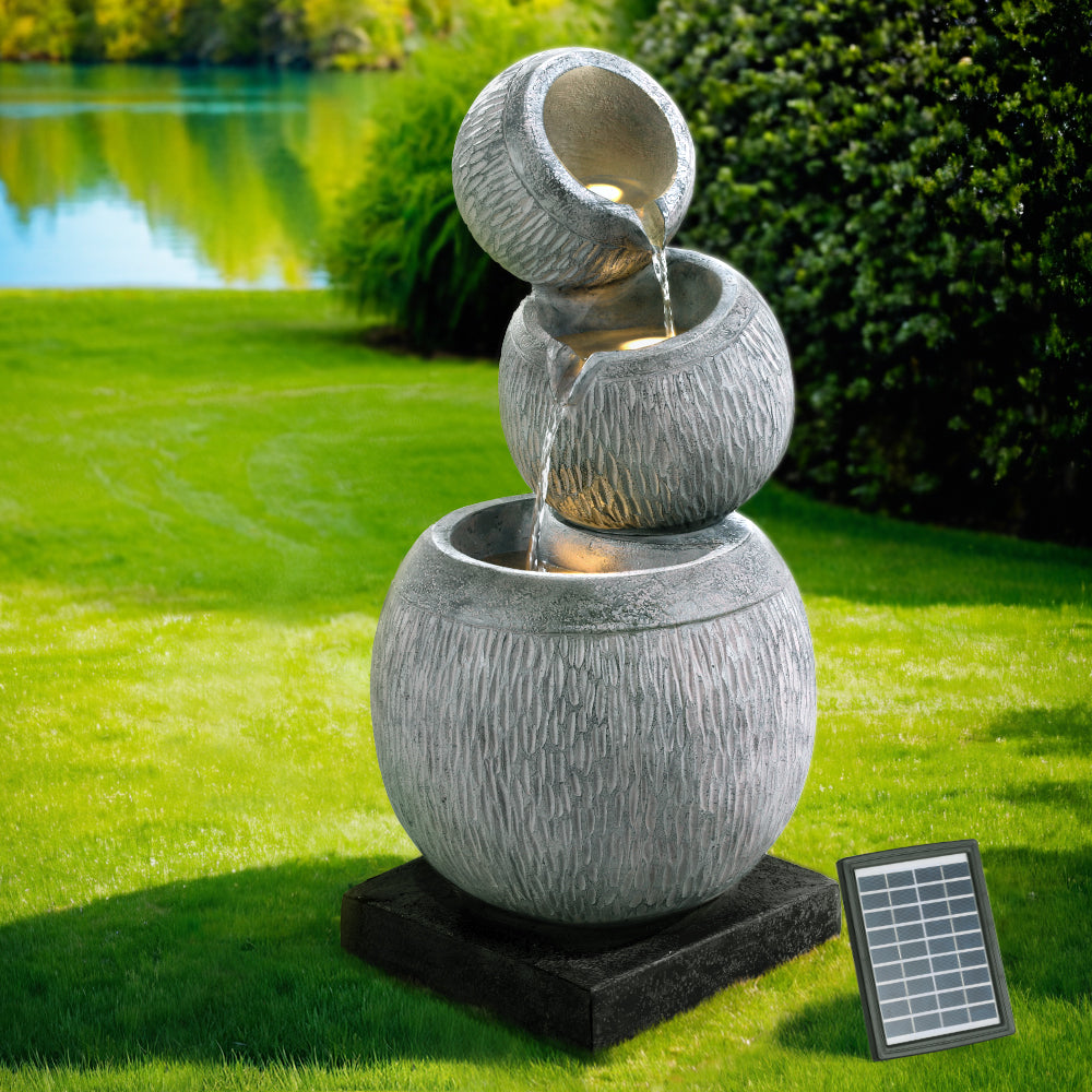 A three-tier bowls cascading water fountain feature with LED lights, made of polyresin, in a grey colour with black base, all in a garden setting on lawn with trees and a lake in the background.