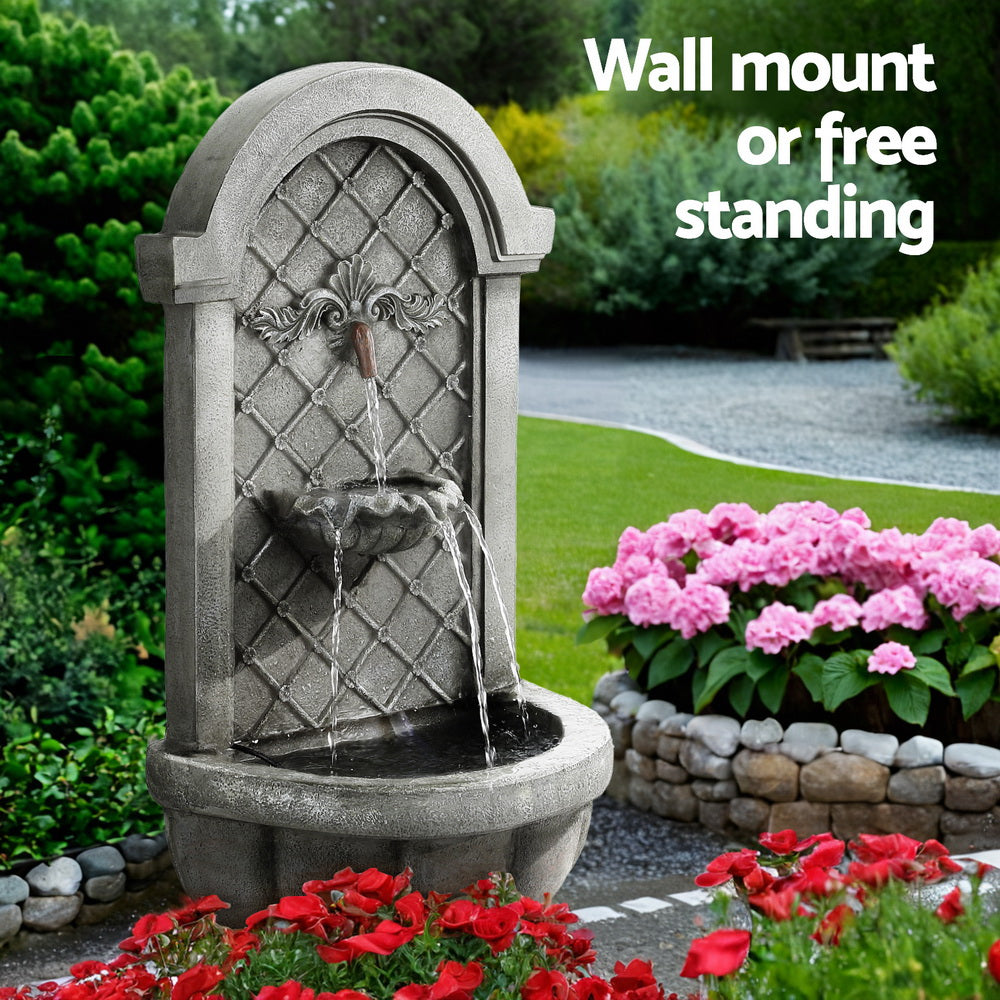 A grey Gardeon solar fountain with a classic cascading design, standing in a pretty flowering rock garden with grass, gravel area with picnic bench and trees in the background.