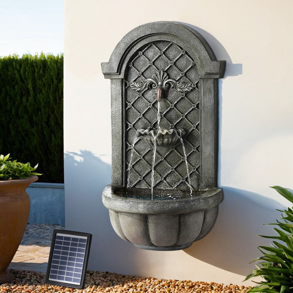 A grey Gardeon solar fountain with a classic cascading design, wall mounted on a white wall with pebbles on the ground below it, a pot plant to the side and a hedge in the background.