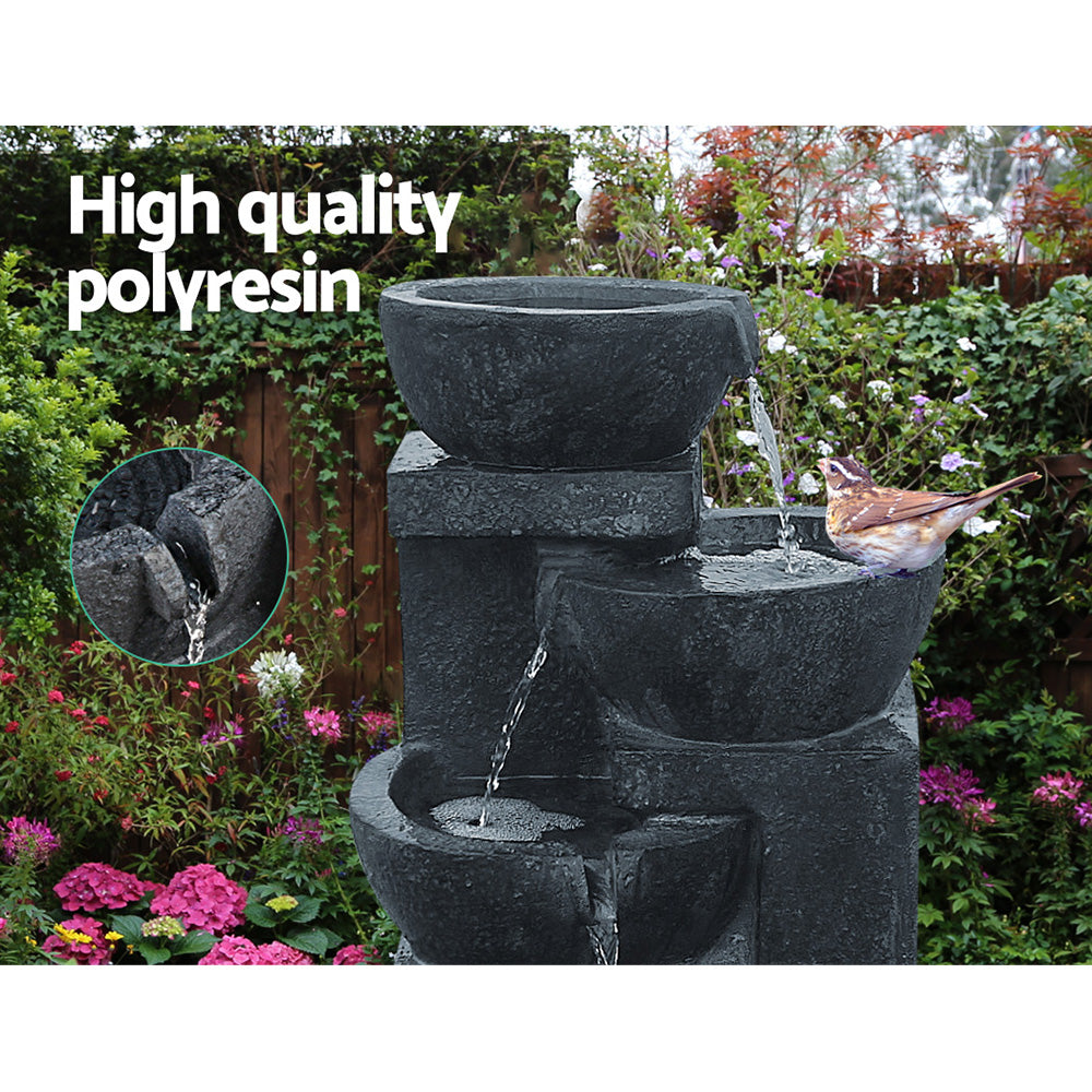 A four-tier cascading water feature with bowls, in blue colour, featuring a bird sitting on the middle bowl, all set in a pretty flowering garden before a wooden fence with trees in the background.