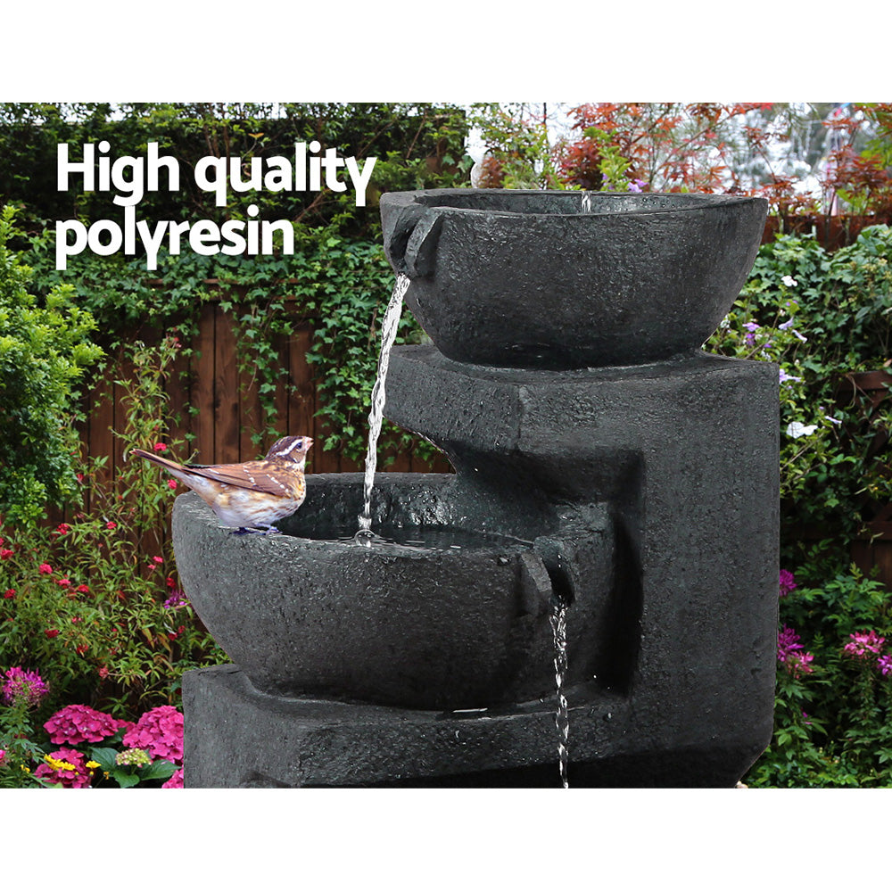 A three-tier cascading water feature with bowls, in blue colour, featuring a bird sitting on the middle bowl, all set in a pretty flowering garden before a wooden fence with flowers and bushes in the background.