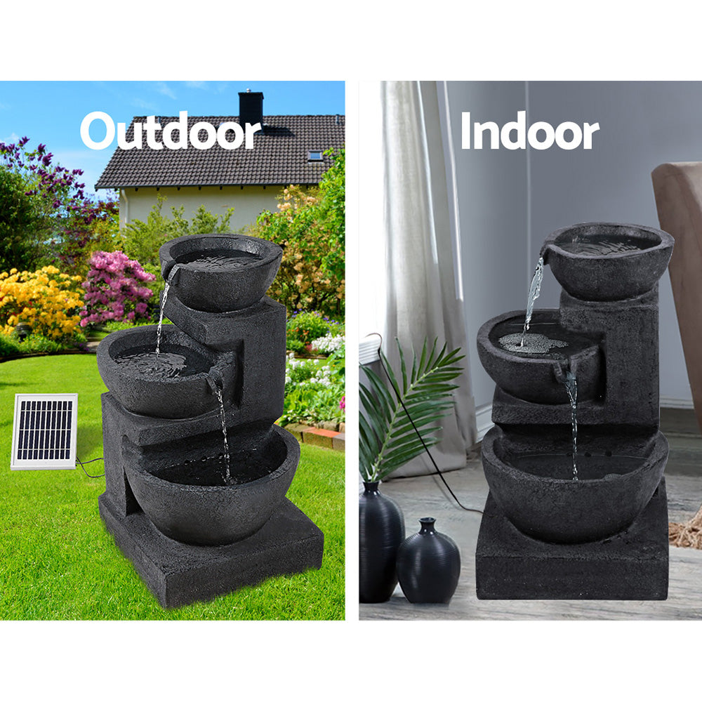 A three-tier blue coloured cascading water feature with bowls, featuring a split photo showing indoor and outdoor use. Outdoor photo is set in a pretty flowering garden on a green lawn with a house in the background and the solar panel standing to the right of the fountain. Indoor photo is near a window with cream curtain and the cord to the solar panel runs outside the window, on a marble floor with a couple of vases standing beside it and a palm leaf in one of them.