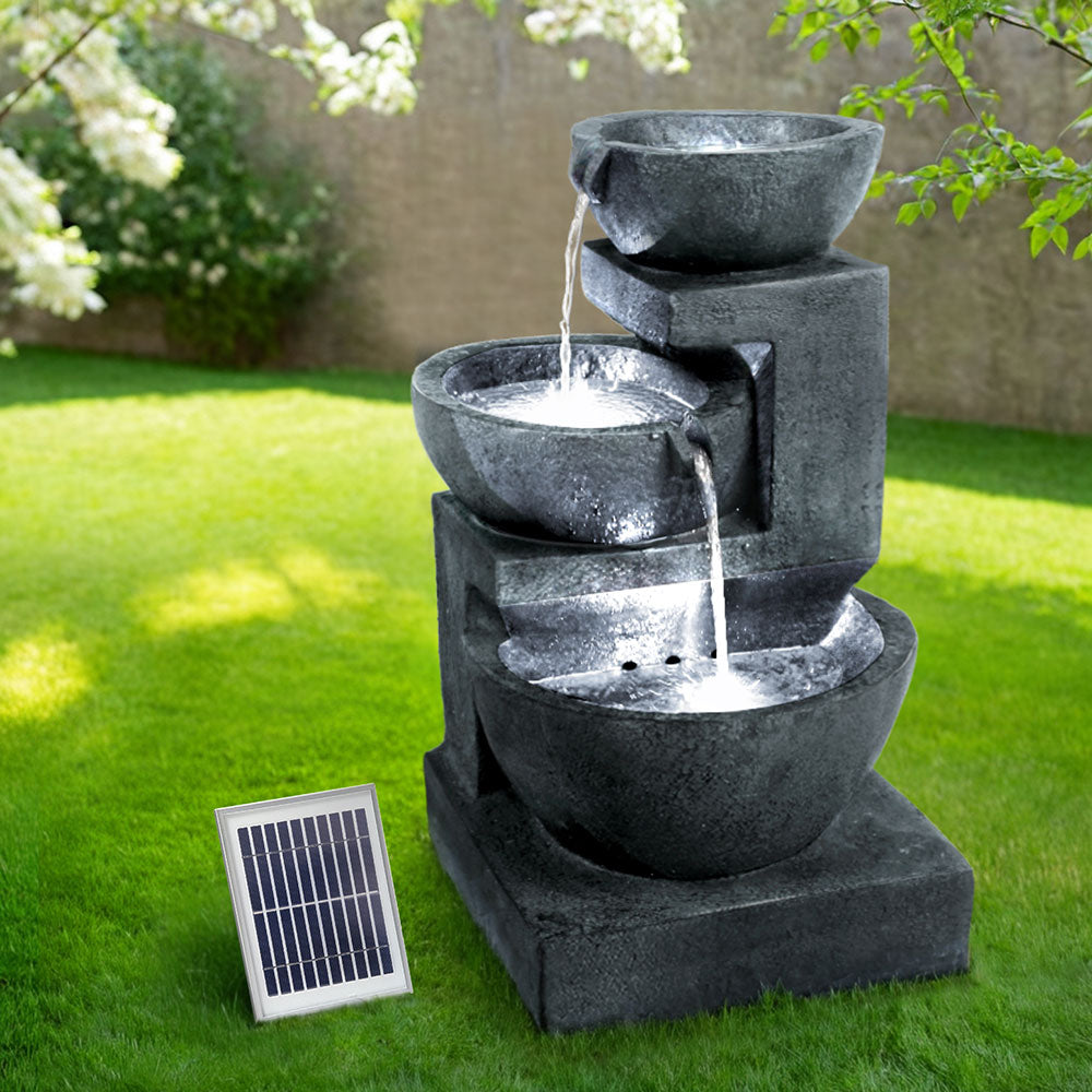 A blue three-tier cascading water feature with bowls, set in a garden before a brick wall on green lawn area with trees in the background.