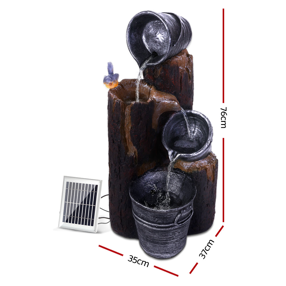 A 3-tier cascading fountain featuring buckets that pour into each other, a bird sitting on a tree stump, a solar panel on the side, and LED lights. The fountain is set against a white backdrop and shows dimensions of the product.