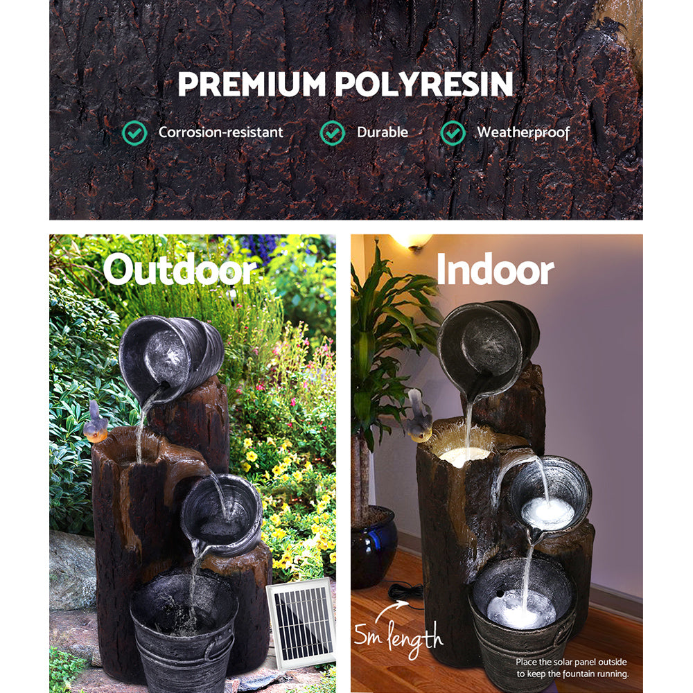 A split image showcasing a 3-tier cascading fountain featuring buckets and tree stump that pour into each other. There is a bird sitting on the tree stump, a solar panel on the side, and LED lights. The fountain shown in Outdoor and Indoor settings. The Outdoor image set in a rockery flowering garden and the Indoor one on a wood floor surface in front of a grey wall with wall light and a pot plant behind it.