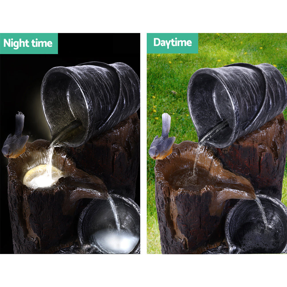 A close up of the top of a 3-tier cascading fountain featuring buckets and tree stump that pour into each other, with a bird sitting on the tree stump. The image is split in two showcasing night and daytime views, the fountain is set against a grass background by day and lit up at night.