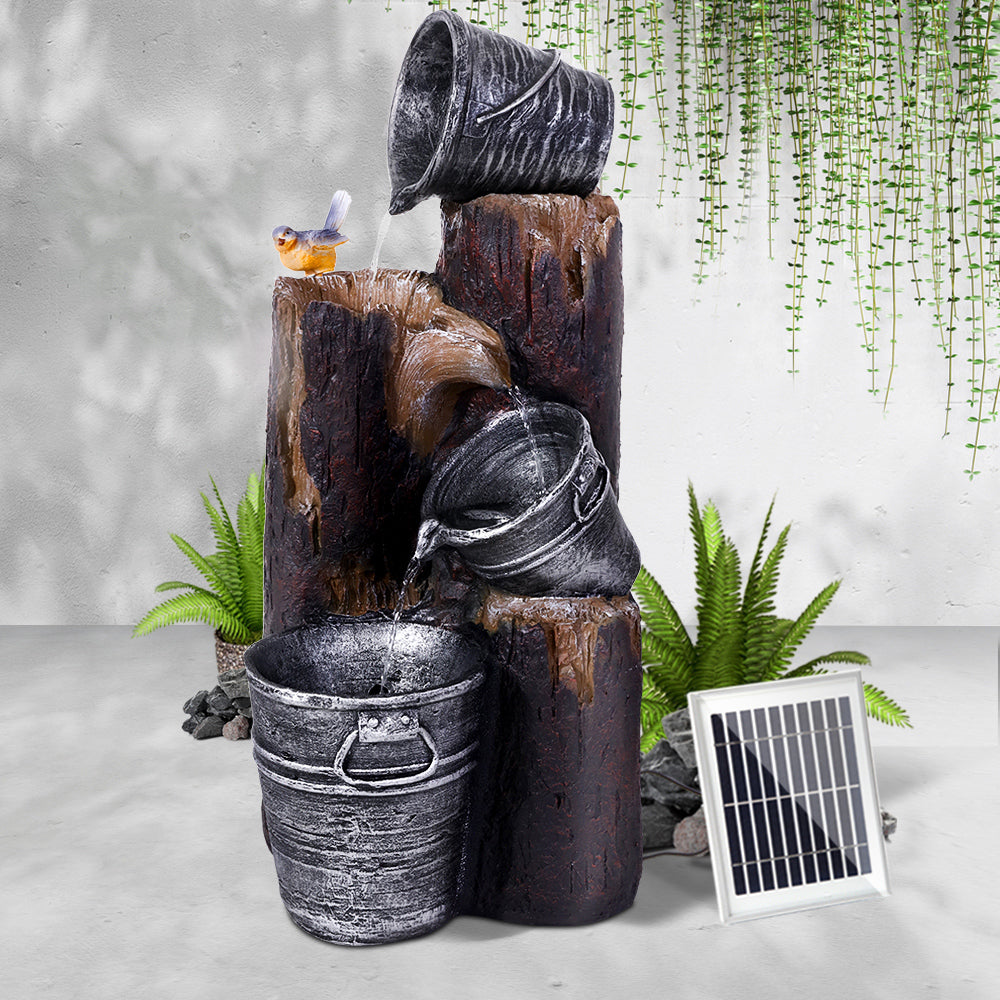 A three-tier cascading water feature with buckets and tree stump, featuring a bird; set in an area in front of a white painted wall on cemented surface with hanging plant in the background.