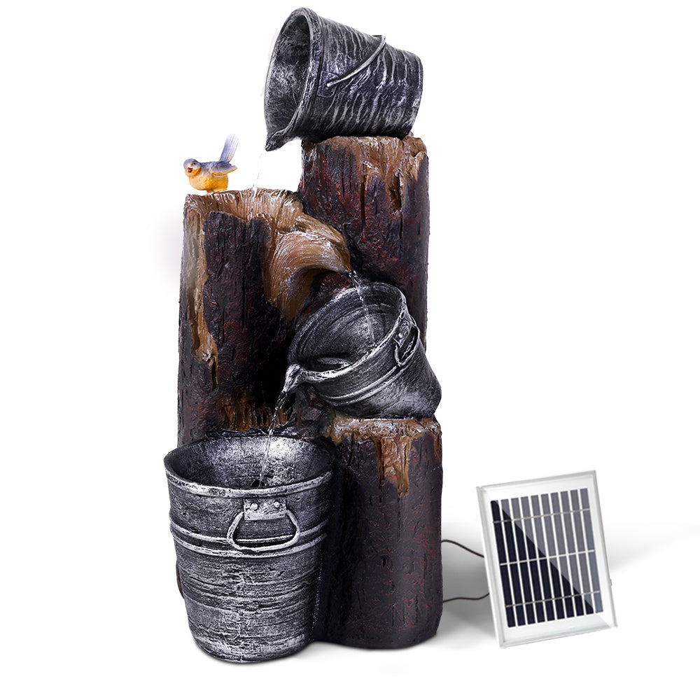 A 3-tier cascading fountain featuring buckets at the top that pours into others, a bird sitting on a tree stump, a solar panel on the side, and LED lights. The fountain is set against a backdrop of plain white.