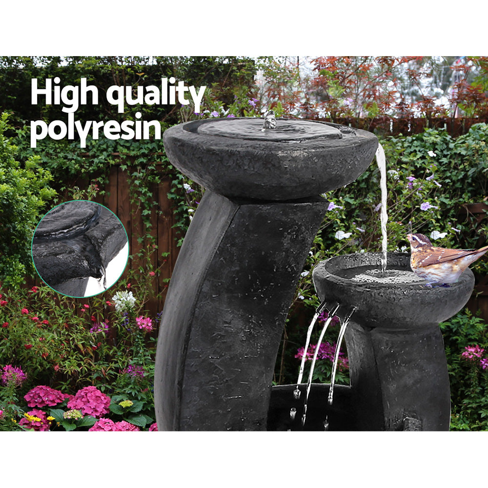 A three-tier cascading water feature with mushroom bowls, in black colour, featuring a bird sitting on the middle bowl, all set in a pretty flowering garden before a wooden fence with flowers and bushes in the background.