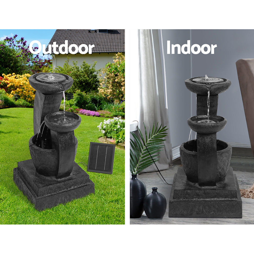 A three-tier black coloured cascading water feature with mushroom bowls, featuring a split photo showing indoor and outdoor use. Outdoor photo is set in a pretty flowering garden on a green lawn with a house in the background and the solar panel standing to the right of the fountain. Indoor photo is near a window with cream curtain and the cord to the solar panel runs outside the window, on a marble floor with a couple of vases standing beside it and a palm leaf in one of them.