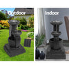 A three-tier black coloured cascading water feature with mushroom bowls, featuring a split photo showing indoor and outdoor use. Outdoor photo is set in a pretty flowering garden on a green lawn with a house in the background and the solar panel standing to the right of the fountain. Indoor photo is near a window with cream curtain and the cord to the solar panel runs outside the window, on a marble floor with a couple of vases standing beside it and a palm leaf in one of them.
