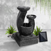 A black three-tier cascading water feature with mushroom bowls, set in an area in front of a white painted wall on cemented surface with hanging plant in the background.