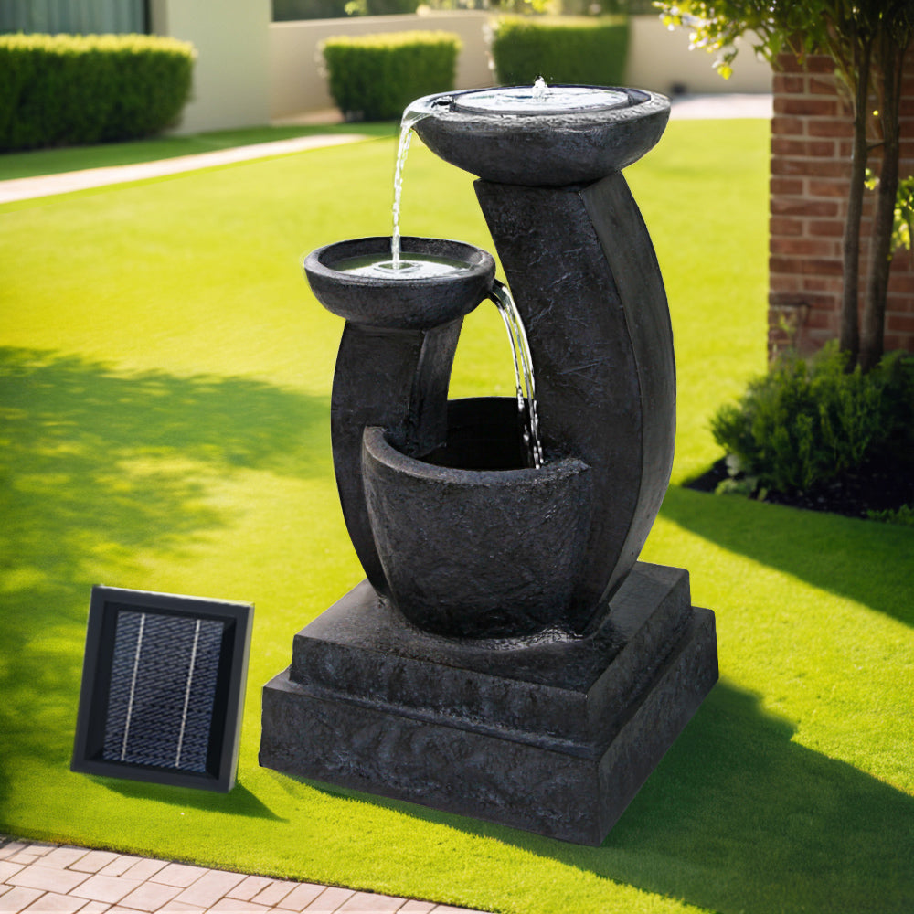 A black three-tier cascading water feature with mushroom bowls, set in a garden near a brick wall on green lawn area with trees and hedges in the background.