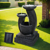 A black three-tier cascading water feature with mushroom bowls, set in a garden near a brick wall on green lawn area with trees and hedges in the background.