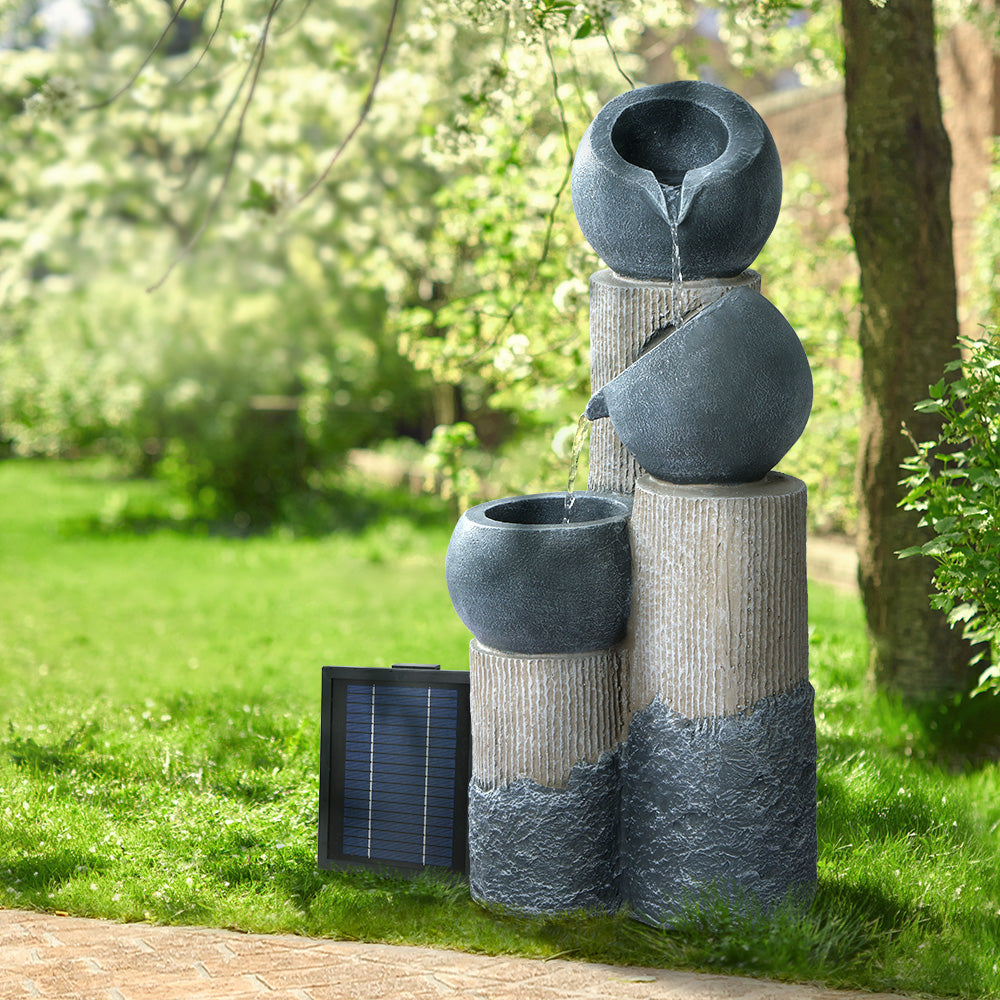 A grey and beige 3-tier cascading water feature with bowls, set in a garden beside a paved walkway, on green lawn area with trees in the background.