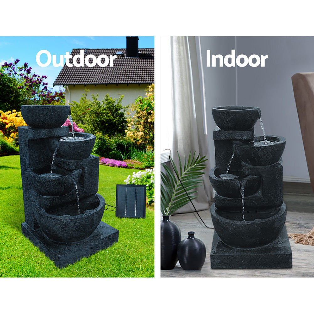 A four-tier blue coloured cascading water feature with bowls, featuring a split photo showing indoor and outdoor use. Outdoor photo is set in a pretty flowering garden on a green lawn with a house in the background and the solar panel standing to the right of the fountain. Intdoor photo is near a window with cream curtain and the cord to the solar panel runs outside the window, on a marble floor with a couple of vases standing beside it and a palm leaf in one of them.