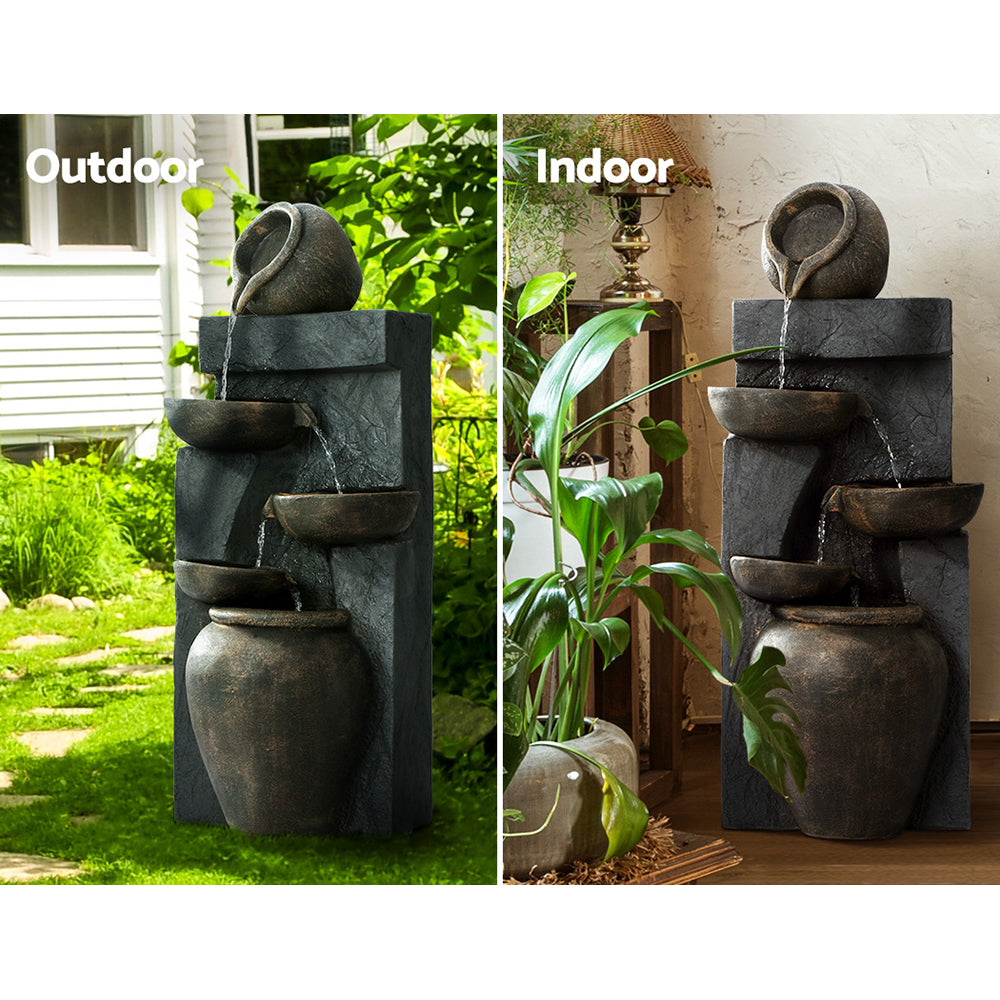 A brown and copper toned, cascading fountain with five-tiered with water flowing down each tier. There are 2 images showcasing indoors and outdoors. Outdoor photo is set on a green lawn with a house and greenery in the background. Indoor photo is near a cream wall, on a wood flooring with a tall lamp table and lamp standing beside it and a couple of pot plants.