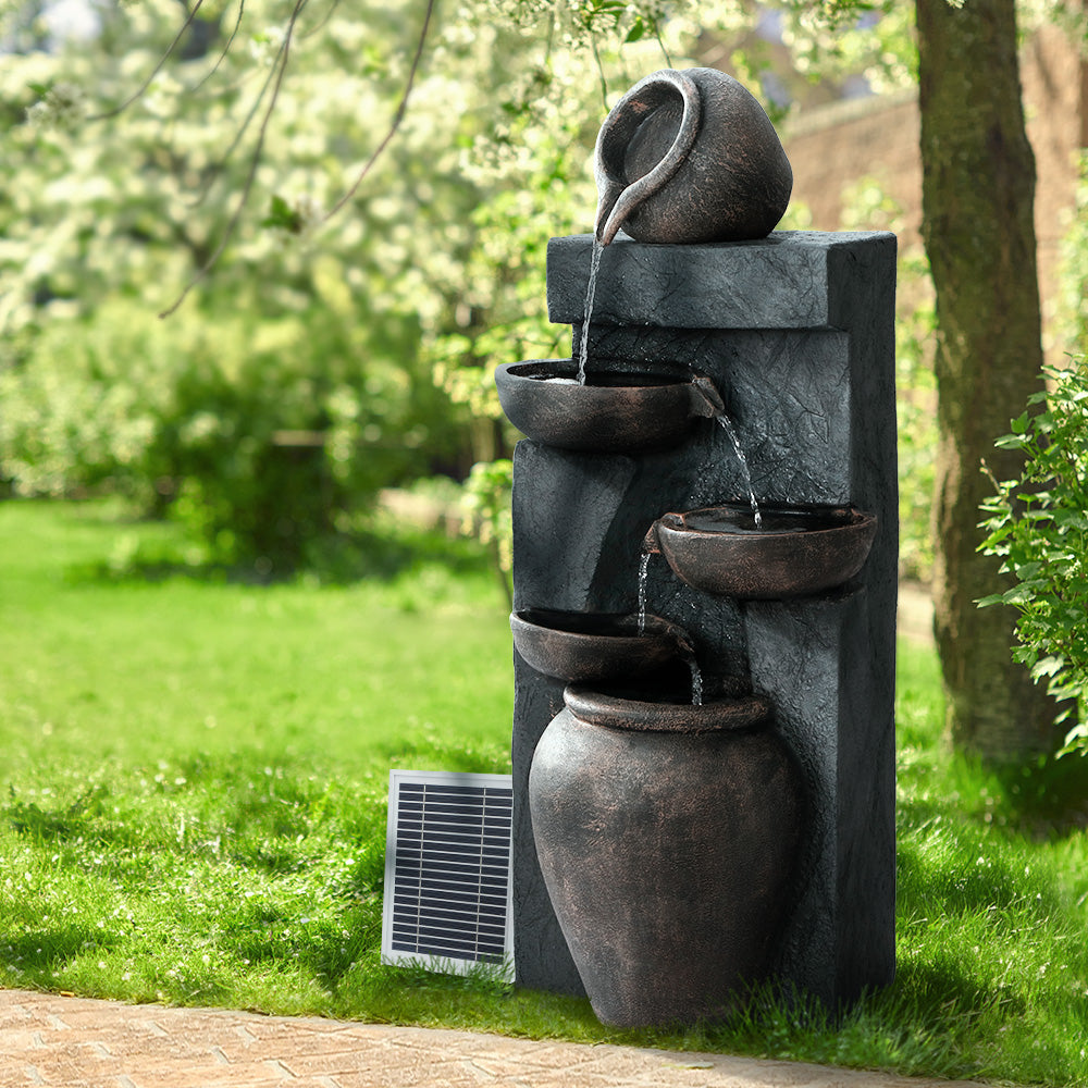 A brown and copper five-tiered cascading water feature with bowls, set in a garden beside a paved walkway, on green lawn area with trees in the background.