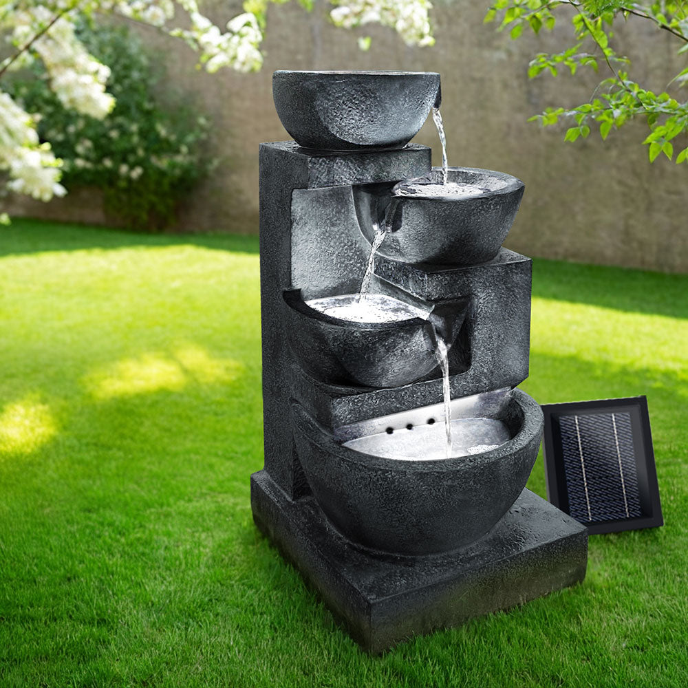 A blue four-tier cascading water feature with bowls, set in a garden before a brick wall on green lawn area with trees in the background.