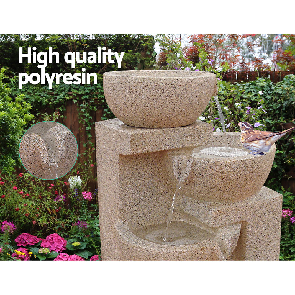 A four-tier cascading water feature with bowls, in blue colour, featuring a bird sitting on the middle bowl, all set in a pretty flowering garden before a wooden fence with trees in the background.