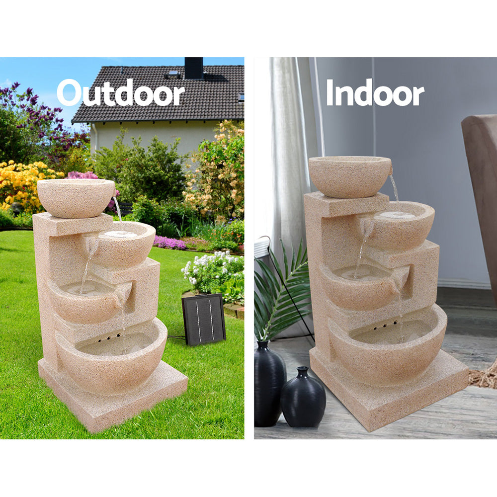 A four-tier sand coloured cascading water feature with bowls, featuring a split photo showing indoor and outdoor use. Outdoor photo is set in a pretty flowering garden on a green lawn with a house in the background and the solar panel standing to the right of the fountain. Indoor photo is near a window with cream curtain and the cord to the solar panel runs outside the window, on a marble floor with a couple of vases standing beside it and a palm leaf in one of them.