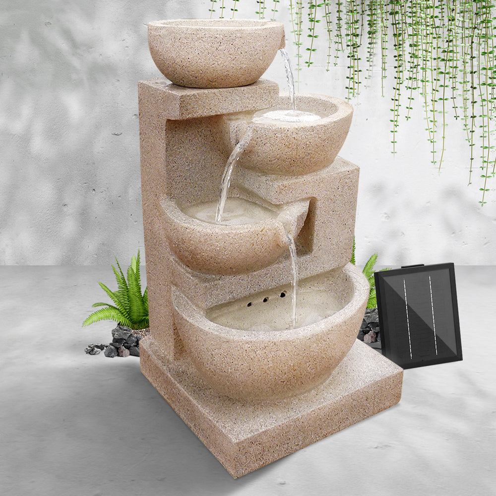 A sand four-tier cascading water feature with bowls, set in an area in front of a white painted wall on cemented surface with hanging plant in the background.