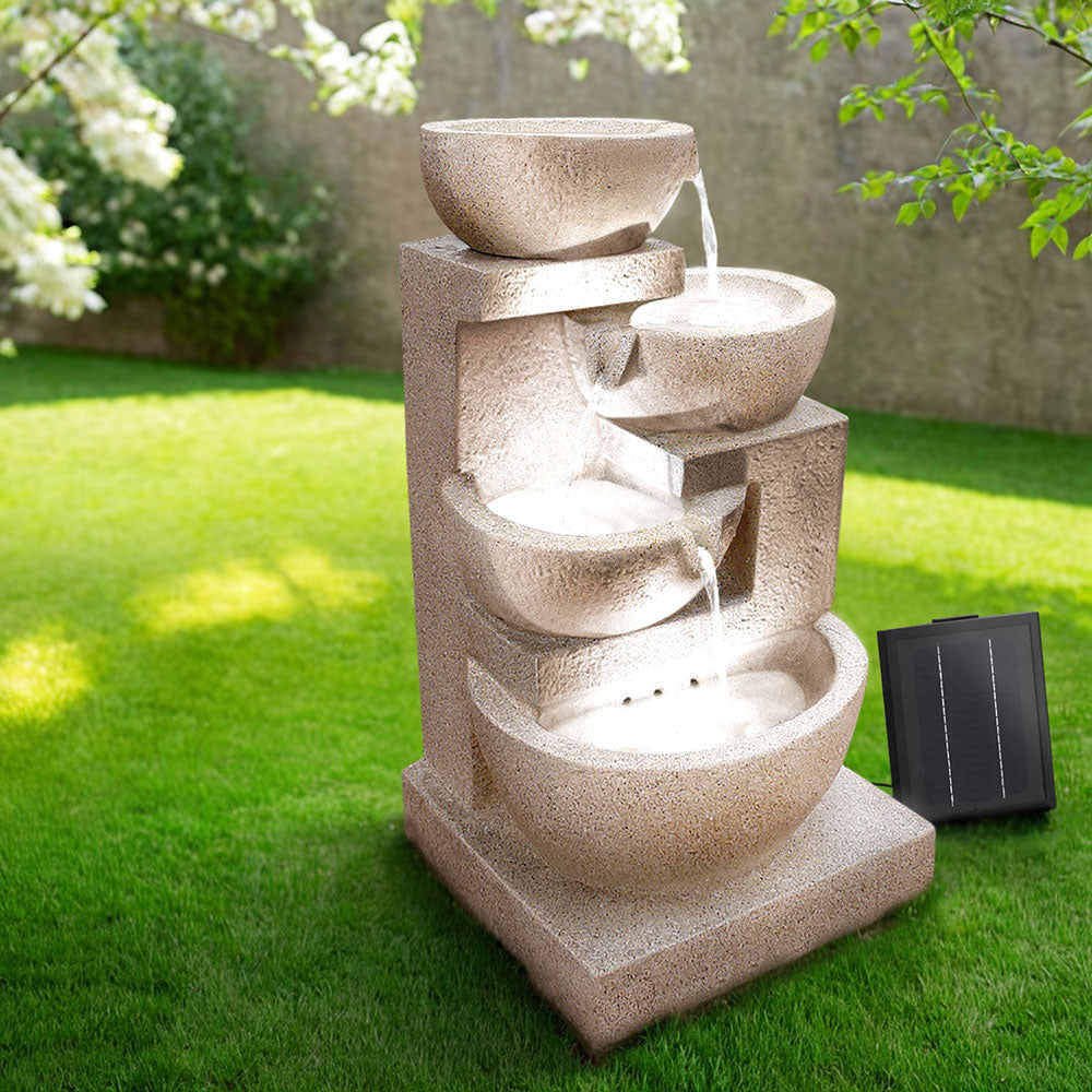 A sand four-tier cascading water feature with bowls, set in a garden before a brick wall on green lawn area with trees in the background.