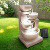 A sand four-tier cascading water feature with bowls, set in a garden before a brick wall on green lawn area with trees in the background.