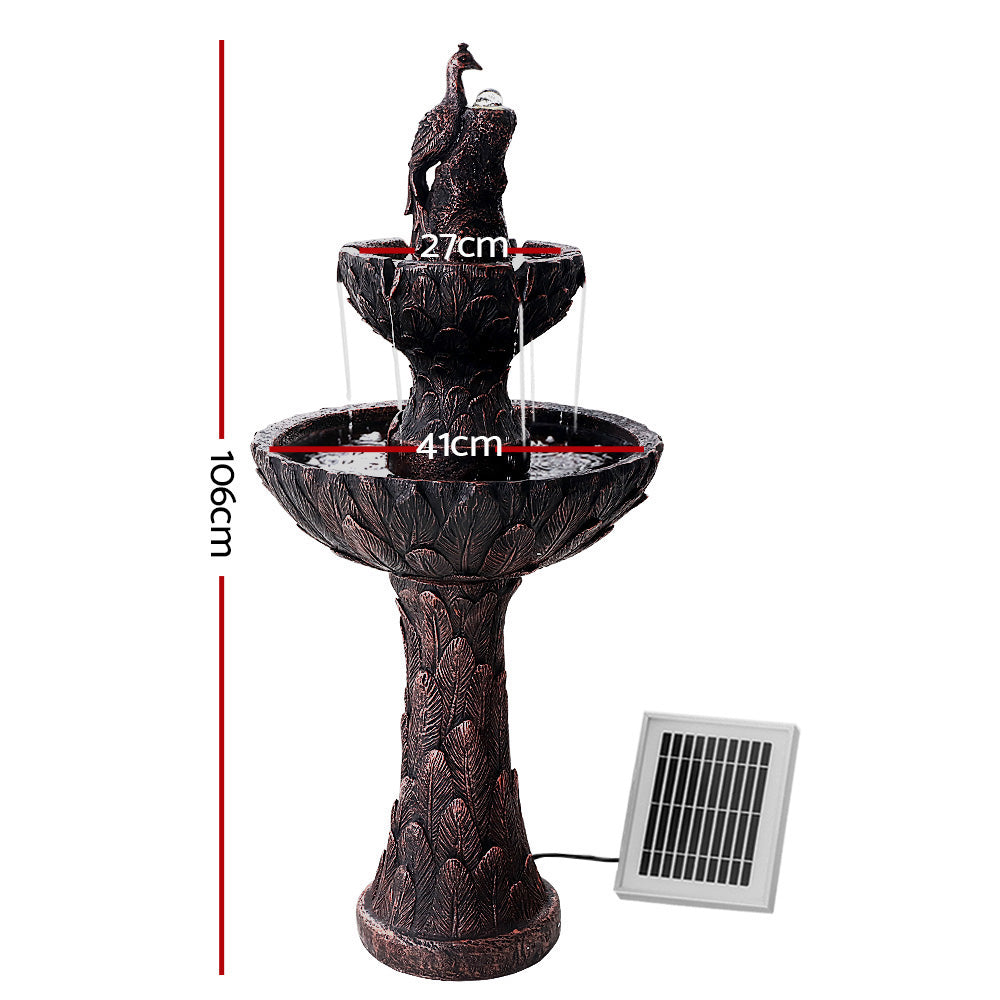 A brown, three-tier solar-powered water fountain featuring a peacock at the top, with a solar panel attached to its base set on a white background showing dimensions of the product.