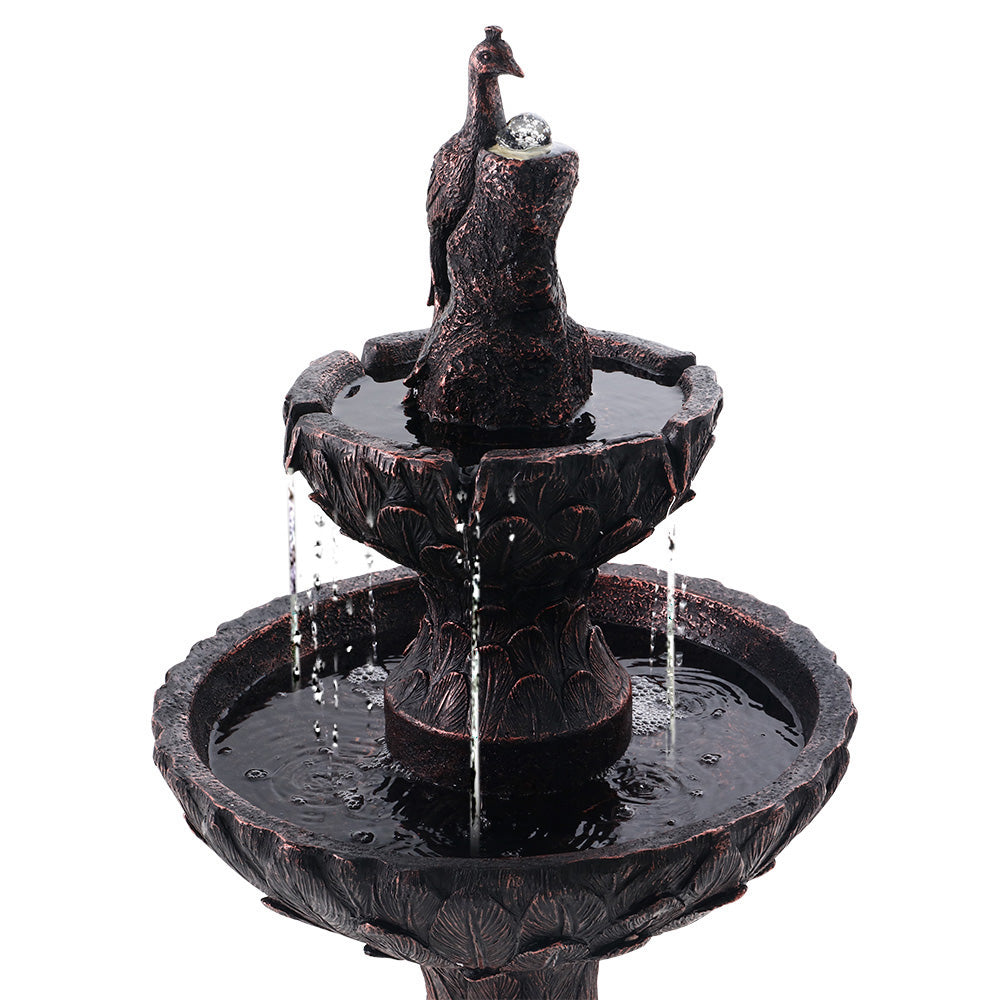 Close up of a brown, three-tier solar-powered water fountain featuring a peacock at the top, set on a white background.