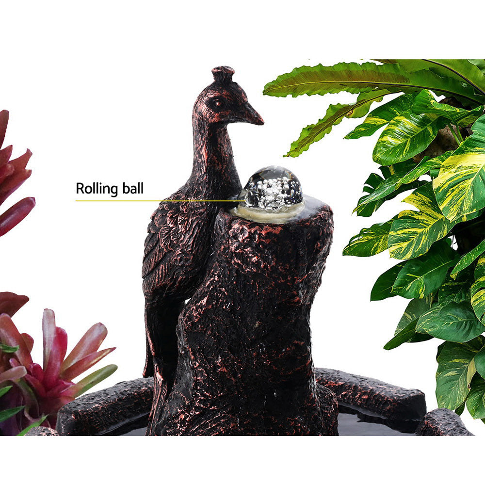 Close up of the peacock and featuring a rolling ball at the top a brown water fountain with plants and on a white background.