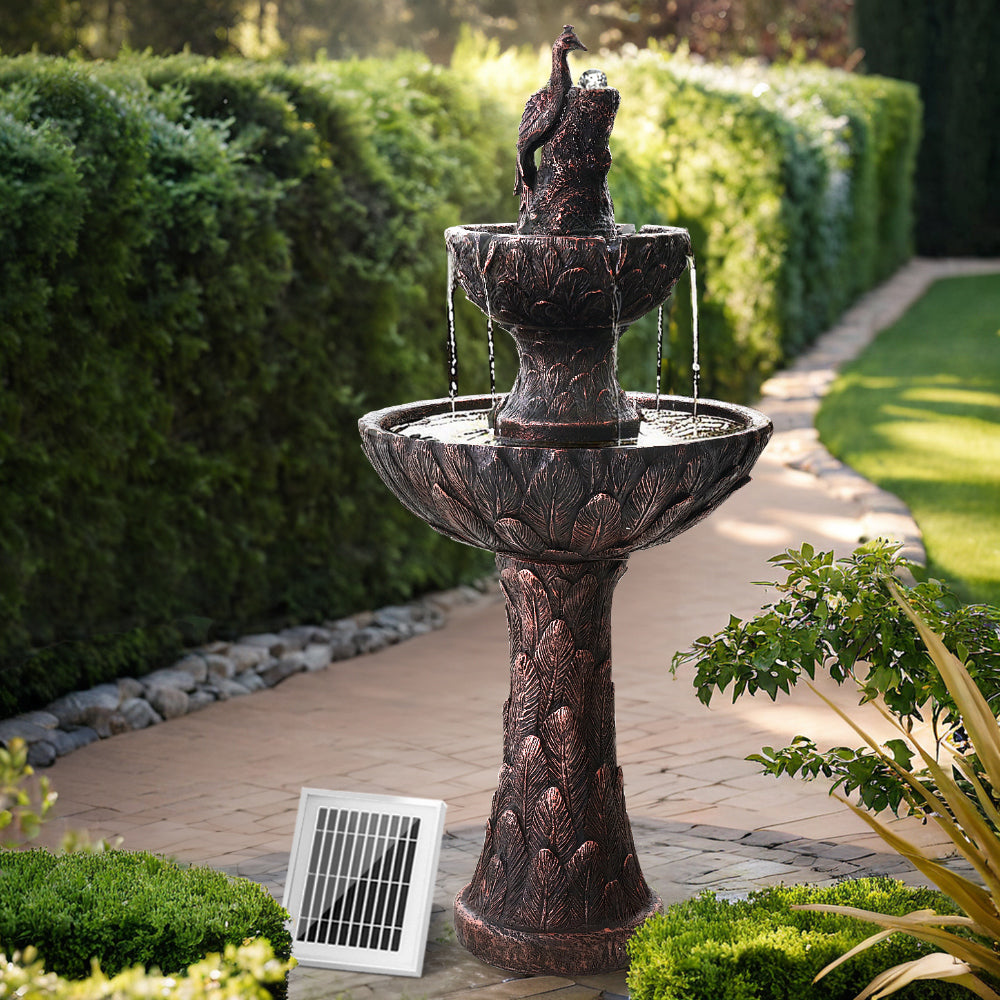 A brown, three-tier solar-powered water fountain featuring a peacock and rolling ball at the top, with a solar panel attached to its base set in a garden with grass, brick pathway, rocks, plants and hedging.