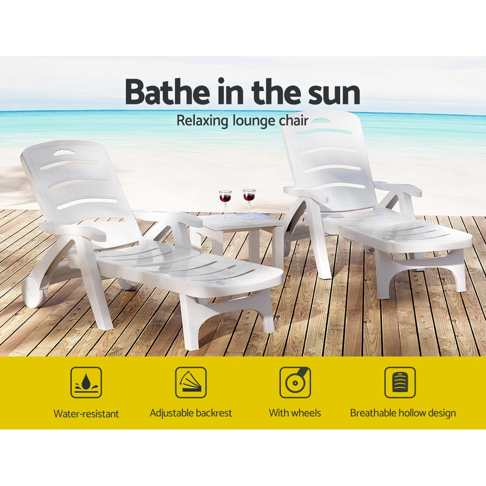 Two white Gardeon folding sun loungers with wheels, in 2 different reclining positions with a small matching table in between them with a couple of glasses of red wine, set on a wood deck with beach in the background.