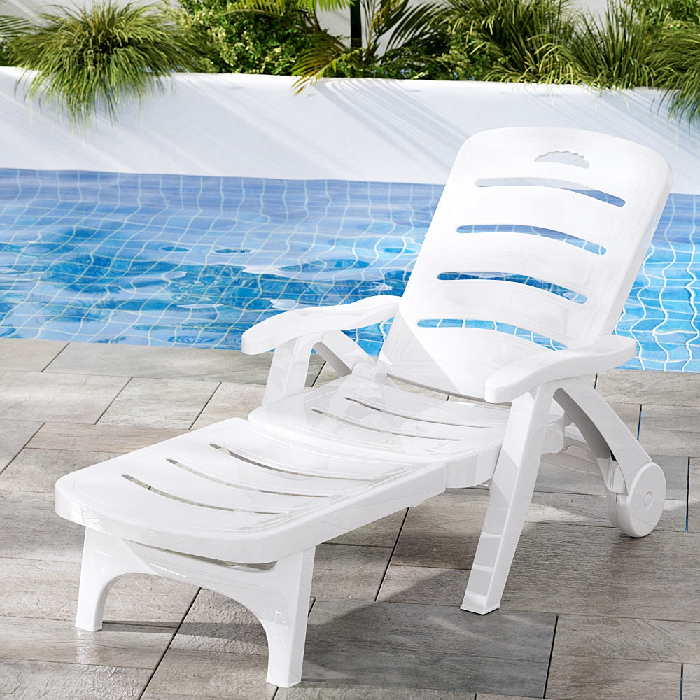 A white Gardeon folding sun lounger with wheels, in a reclined position on a tiled surface in front of a pool with planter boxes and plants on the other side of the pool.