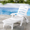 A white Gardeon folding sun lounger with wheels, in a reclined position on a tiled surface in front of a pool with planter boxes and plants on the other side of the pool.