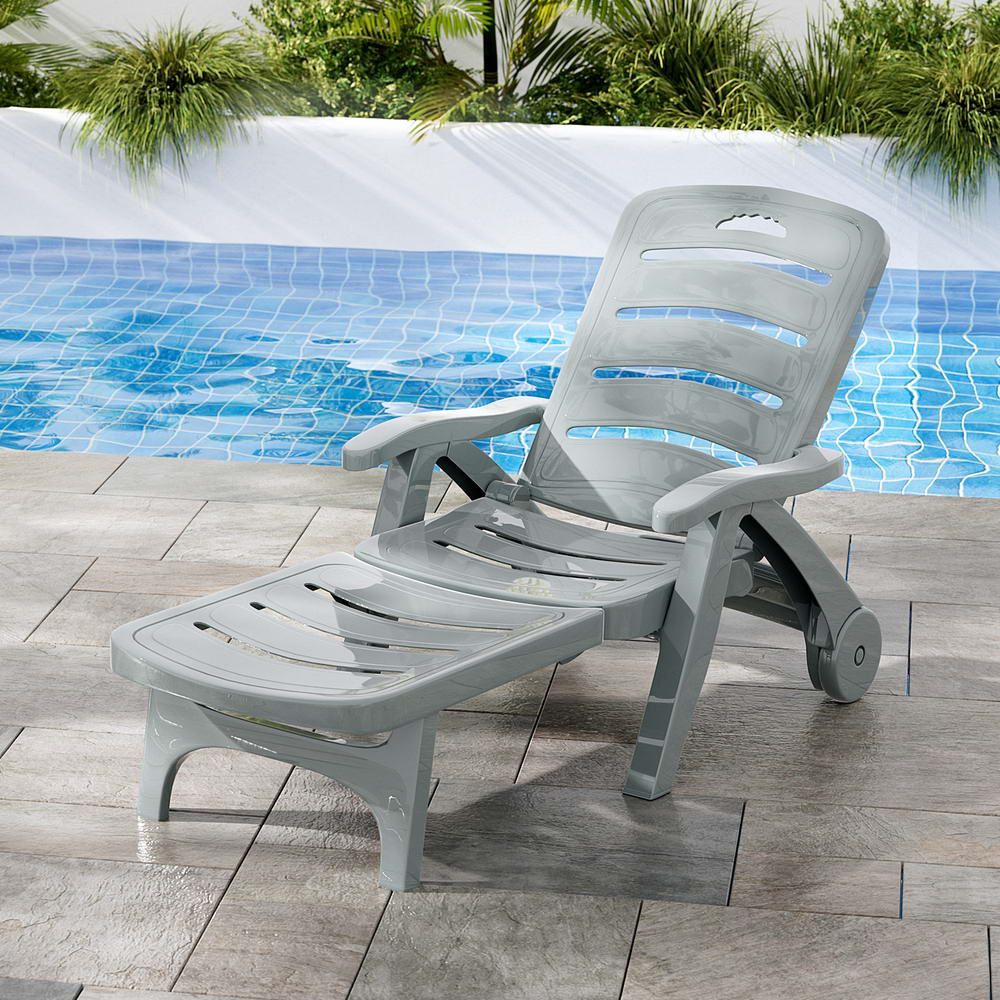 A grey Gardeon folding sun lounger with wheels, in a reclined position on a tiled surface in front of a pool with planter boxes and plants on the other side of the pool.