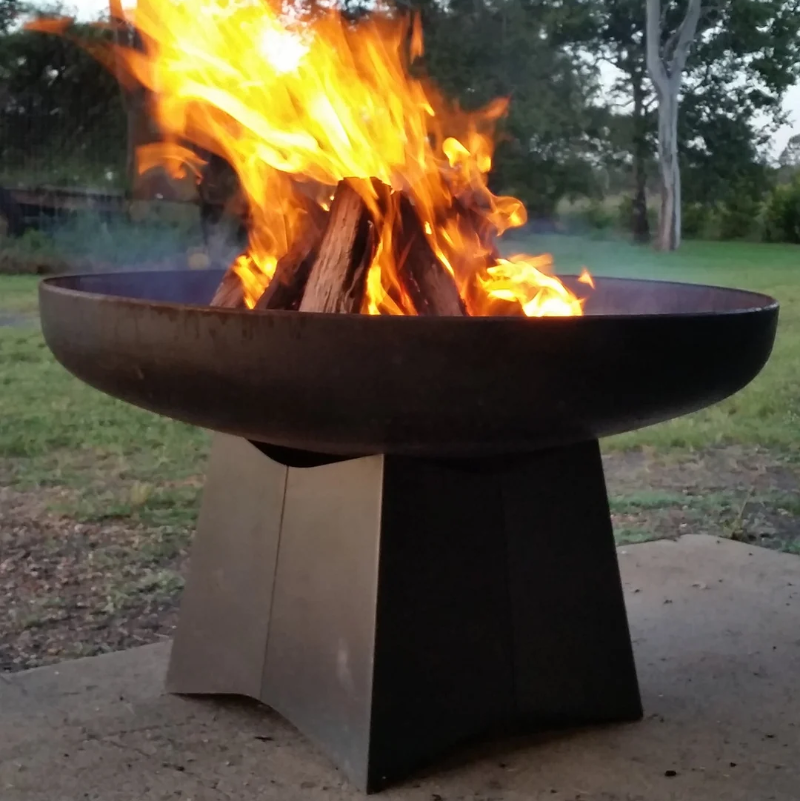 Yagoona Goanna Outdoor Fire Pit Australia with a wood fire burning nicely, sitting on a cement slab within a grassy area with trees in the background.