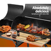 The black Grillz 2-in-1 offset charcoal smoker with a chrome-coated steel BBQ grill on top cooking steaks and chicken pieces and a side firebox with a whole chicken roasting in it. Its large front shelf has condiments and cooked chicken pieces, set on a white background.