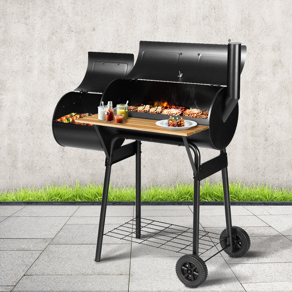 A black Grillz 2-in-1 offset charcoal smoker with a chrome-coated steel BBQ grill on top and a side firebox, both with steaks, chicken pieces and kebabs cooking. It has a large front shelf with condiments and a plate of cooked kebabs and is set on grey paving tiles with grass and grey wall in the backdrop.