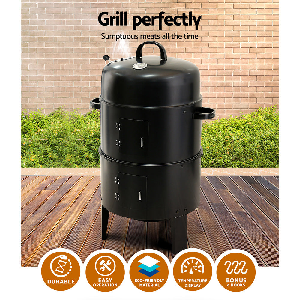 A black 3-in-1 charcoal BBQ smoker with a lid, side doors, and a front-mounted temperature gauge, set on a wooden deck with hedging and brick wall in the backdrop.