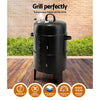 A black 3-in-1 charcoal BBQ smoker with a lid, side doors, and a front-mounted temperature gauge, set on a wooden deck with hedging and brick wall in the backdrop.
