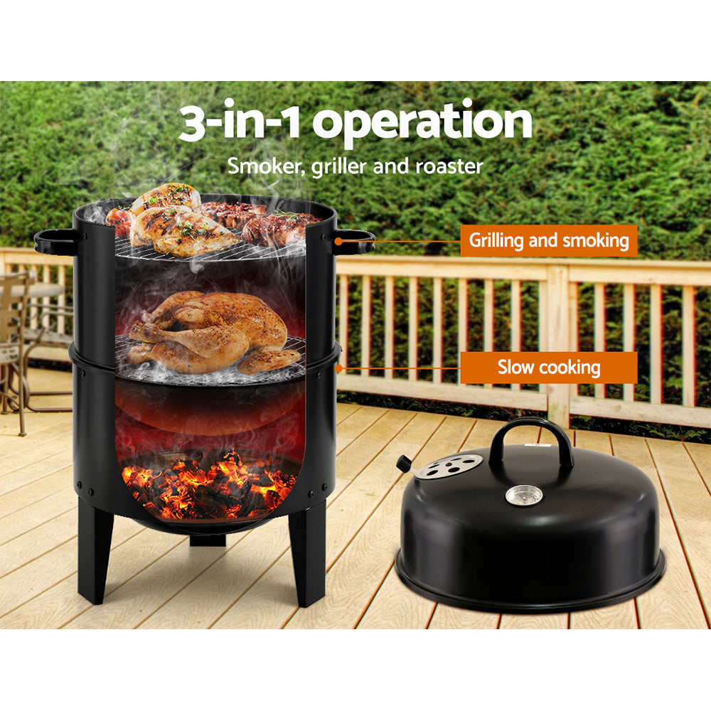 A black 3-in-1 charcoal BBQ smoker with a lid beside it, showcasing the inside where it has a whole chicken roasting in the middle section and chicken breasts, steaks and tomatoes grilling and smoking on the top grill; set on a wooden deck with wooden fencing and trees in the background.