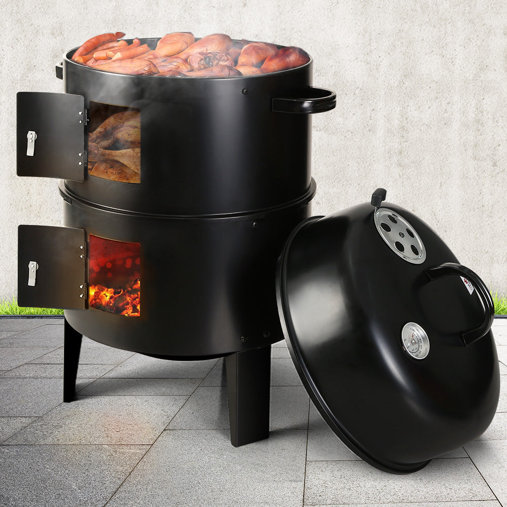 A black 3-in-1 charcoal BBQ smoker with its lid leaning against the side of it, side doors are open showing inside flames at bottom, a roasting chicken in the middle and chicken pieces grilling and smoking on top grill, on a tiled outdoor area with grass and grey wall in the backdrop.