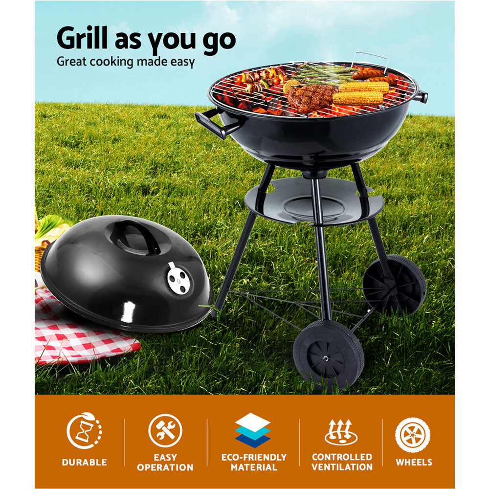 A black portable BBQ smoker with a chrome-coated steel grill, with a steak, kebabs, corn cobs and asparagus cooking, set on a grass with picnic rug and basket.