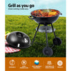 A black portable BBQ smoker with a chrome-coated steel grill, with a steak, kebabs, corn cobs and asparagus cooking, set on a grass with picnic rug and basket.