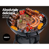 A black portable BBQ smoker with a chrome-coated steel grill, with steaks, ribs, sausages and vegetables cooking, set against a dark background.