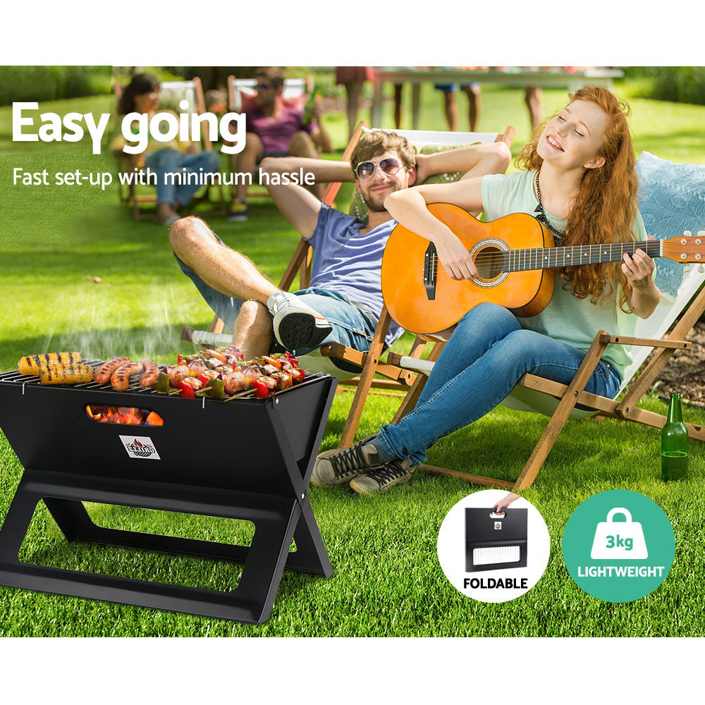 A black Grillz Foldable BBQ Grill Charcoal Smoker cooking sausages, kebabs and corn cobs, set on a grass area with two people in deck chairs, a young man relaxed and a young woman playing guitar, more people can be seen in the background. 