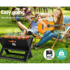 A black Grillz Foldable BBQ Grill Charcoal Smoker cooking sausages, kebabs and corn cobs, set on a grass area with two people in deck chairs, a young man relaxed and a young woman playing guitar, more people can be seen in the background. 