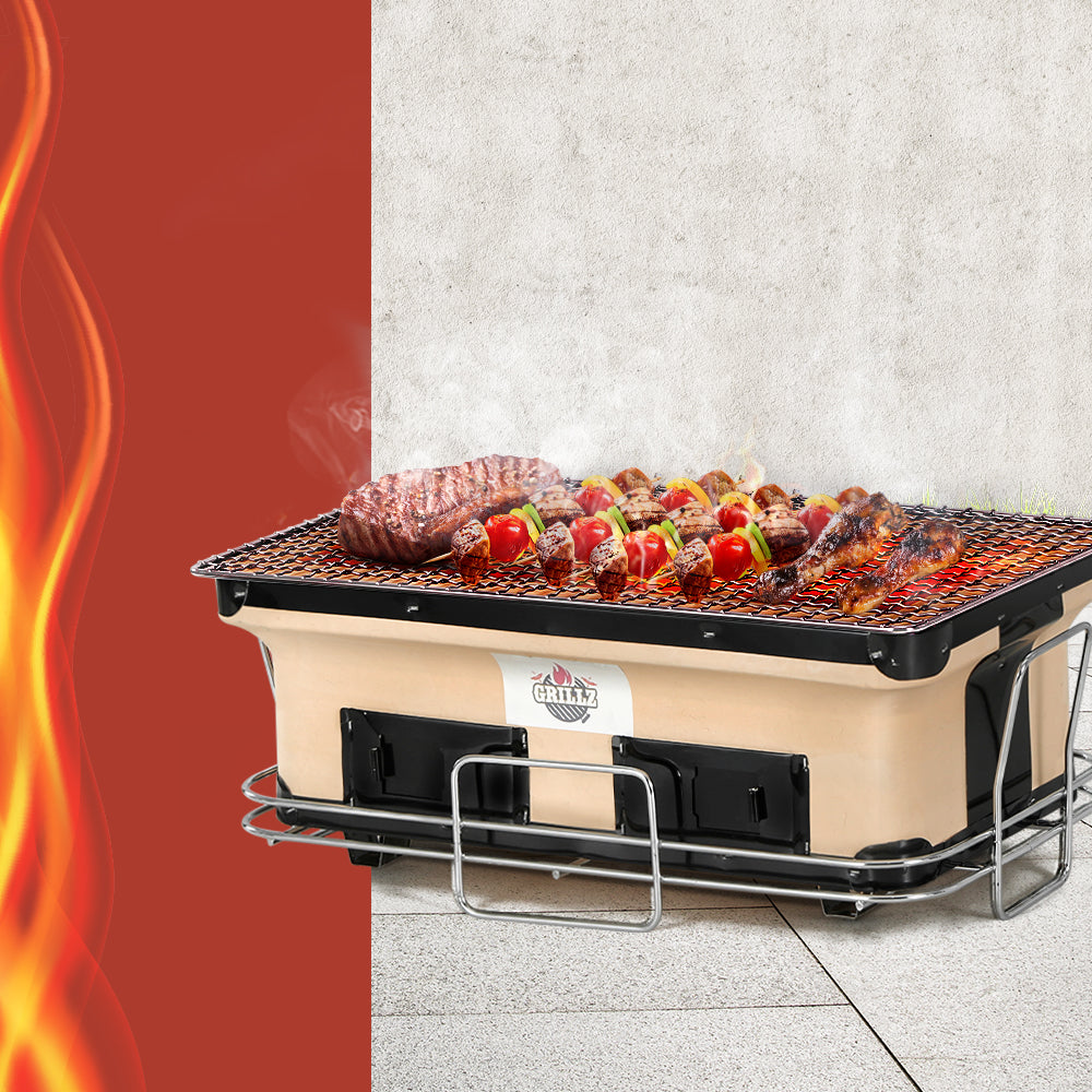 Grillz BBQ Grill Tabletop Charcoal Smoker with setak, skerwered kebabs and chicken drumsticks cooking, set on a tiled floor with painted wall in the background.